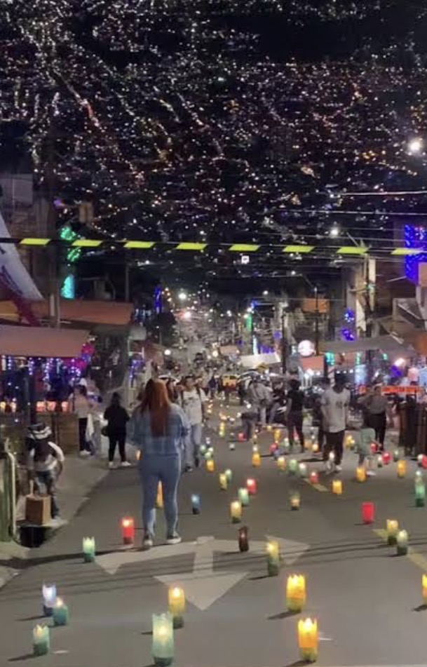Calle de barrio cerrada la noche de las velitas en Medellín, con faroles de colores encendidos sobre el asfalto y la ladera de la ciudad iluminada al fondo