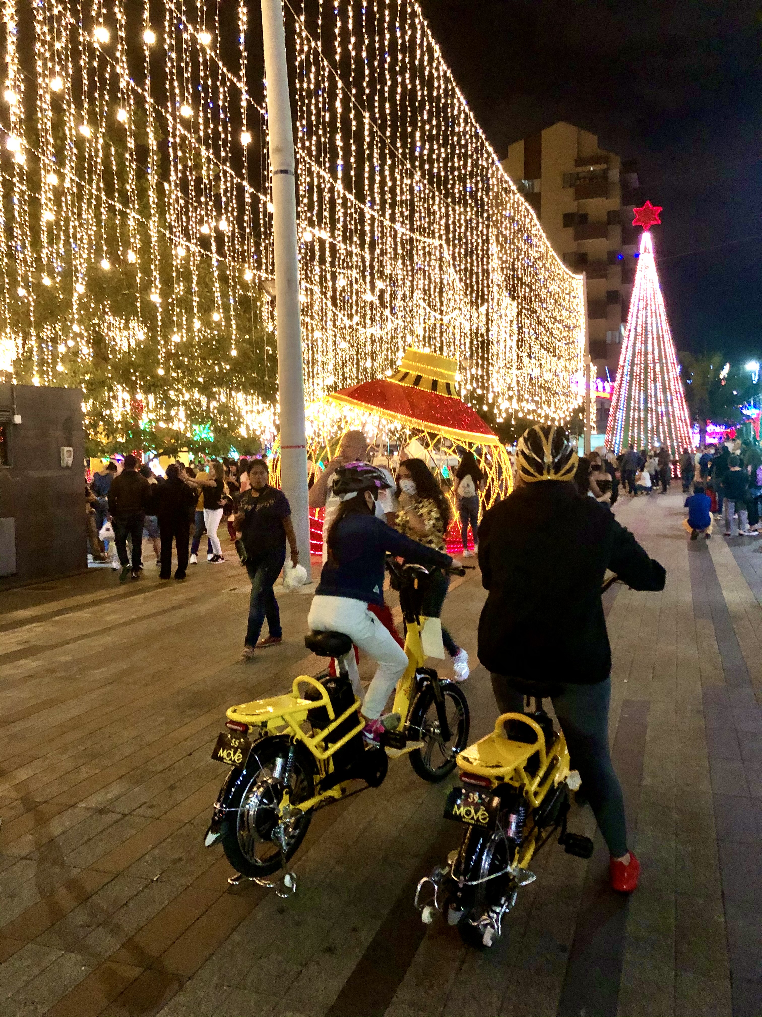 Besucher schieben ihre MOVE-E-Bikes unter einem Vorhang aus Weihnachtslichtern in Medellín, im Hintergrund ein beleuchteter Weihnachtsbaum